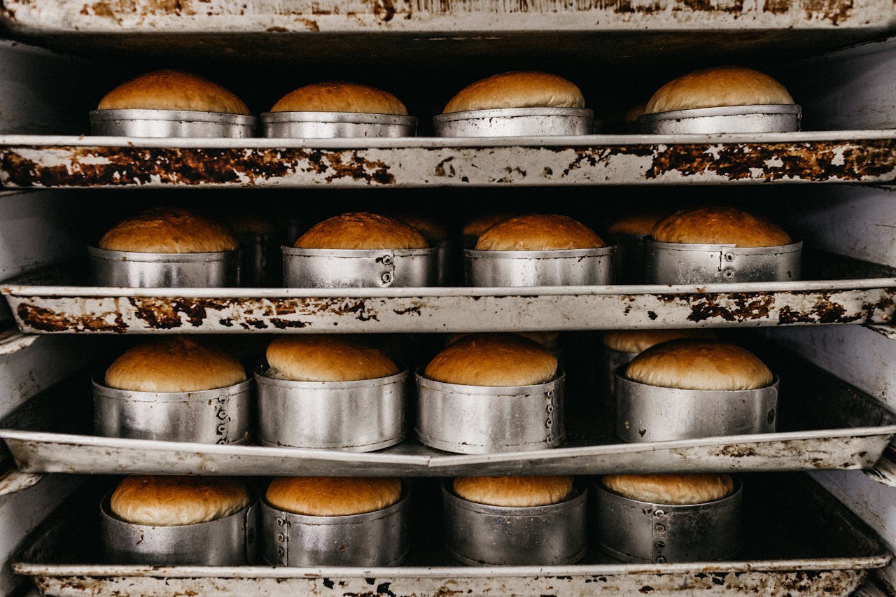 hero-img-02 Rows of golden bread rolls in metal trays, showcasing industrial baking setup.